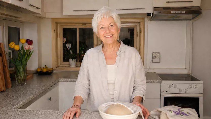 Elfriede hält in ihrer hellen Küche kurz inne, eine Hand an der Arbeitsfläche, und wirkt gesammelt und bei sich.